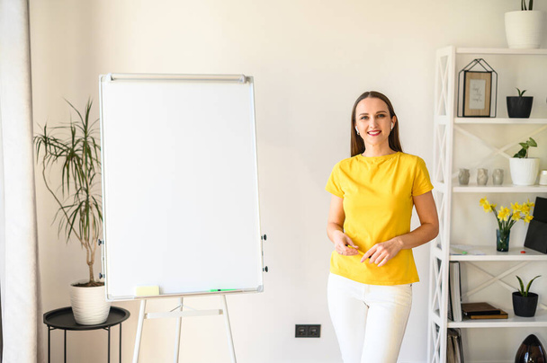 Online učitel, učitel, mentor. Sebevědomá mladá žena ve žlutém tričku stojí v jasné místnosti poblíž prázdné flip chart a usmívá - Fotografie, Obrázek