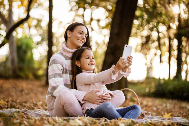 Anya és lánya okostelefont használ ősszel a parkban. Szelfit csinálnak.. - Fotó, kép