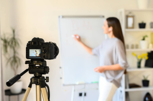 Educational video blogging, online training. A young woman is recording videos on camera. She stands near a flipchart board, points at her and explains something. Focus on screen - Fotografie, Obrázek