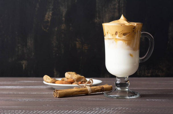Dalgona Coffee Iced, a Korean fashionable whipped black instant coffee with milk, on a brown wooden background. On a white plate with dried figs, almonds, cinnamon sticks. Copy space. Side view. - Foto, Imagem