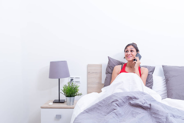 Szczęśliwa młoda kobieta w łóżku uśmiechnięta podczas słuchania przez telefon komórkowy. - Zdjęcie, obraz