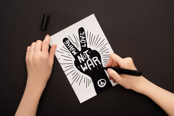 cropped view of woman holding marker near white paper with hand drawing and make love not war lettering on black background - Фото, зображення