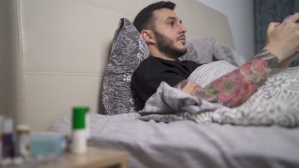 Soustředěný muž hrající videohry v posteli. Noční stolek s léky a pilulkami vedle jeho postele - Záběry, video