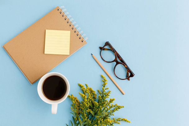 Blank brown vintage notebook, leaves, yellow flowers, diary, cup of coffee, paper clips, eyeglasses on colorful blue table. Stylish minimalistic workplace concept. Top view with copy space. - Photo, Image