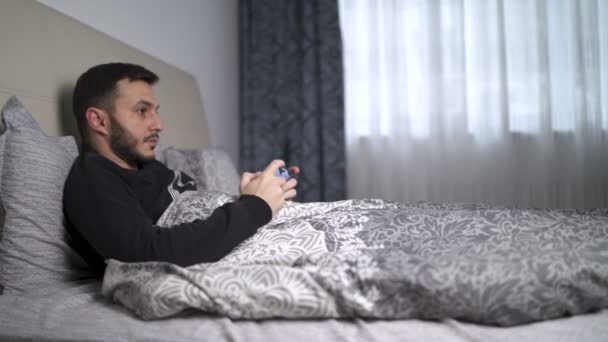 Kwarantanna młody człowiek gra w gry wideo w łóżku za pomocą kontrolera - Materiał filmowy, wideo