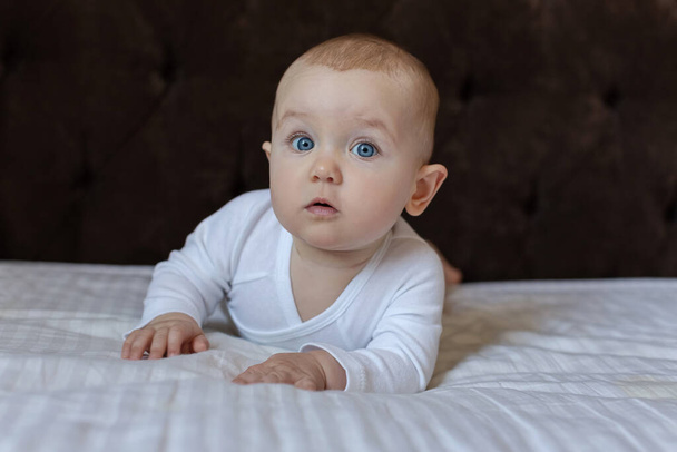 Cute baby in a white light bodysuit on the bed at home looking at the camera. Portrait of a cute baby lying down on a blanket. Baby girl smiling. Portrait of a crawling baby on the bed in the parents room on the big bed. - Zdjęcie, obraz