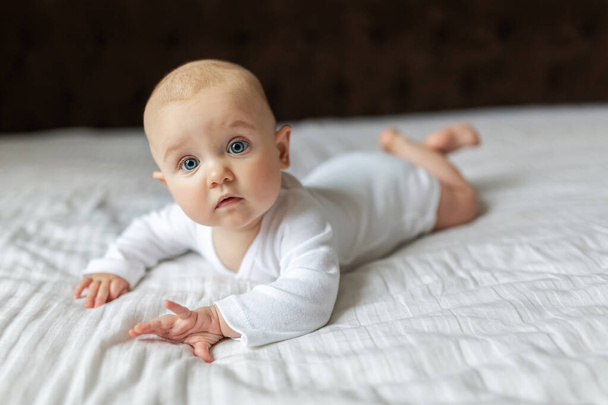 Cute baby in a white light bodysuit on the bed at home looking at the camera. Portrait of a cute baby lying down on a blanket. Baby girl smiling. Portrait of a crawling baby on the bed in the parents room on the big bed. - Photo, image