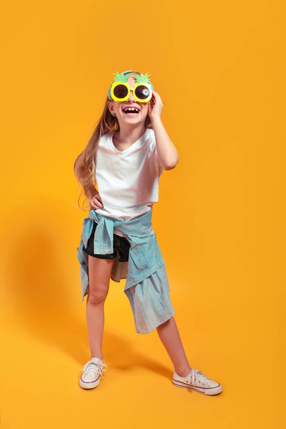 Menina sorridente posando nos óculos de sol engraçados e olhando para a câmera no fundo do estúdio amarelo
 - Foto, Imagem