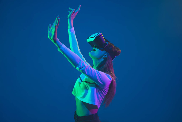 Caucasian womans portrait isolated on blue studio background in pink-green neon light, stylish and beautiful, playing with VR-headset - Photo, image