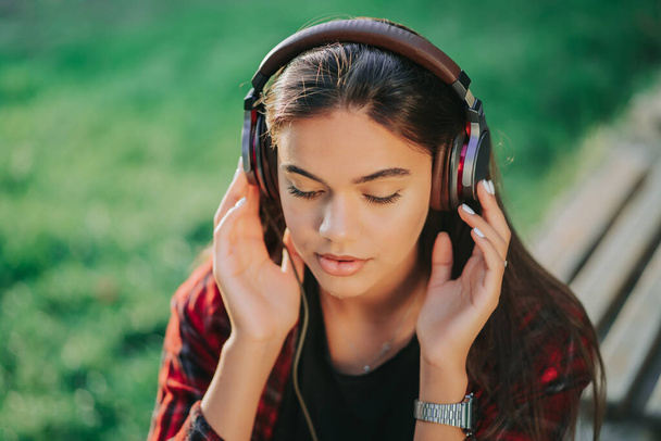 Cute girls face with headphones. Young woman enjoys music while sitting in green park. Concept of youth, freedom, technology - Photo, Image