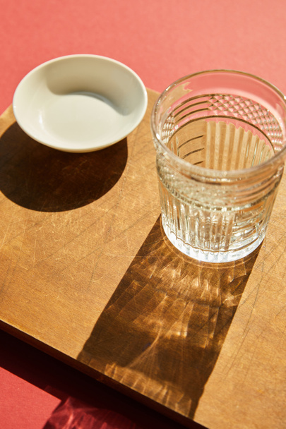 faceted empty glass and white bowl on wooden board in sunlight - Photo, Image
