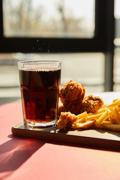 crispy deep fried chicken and french fries served on wooden cutting board with soda in sunlight near window - Foto, Bild