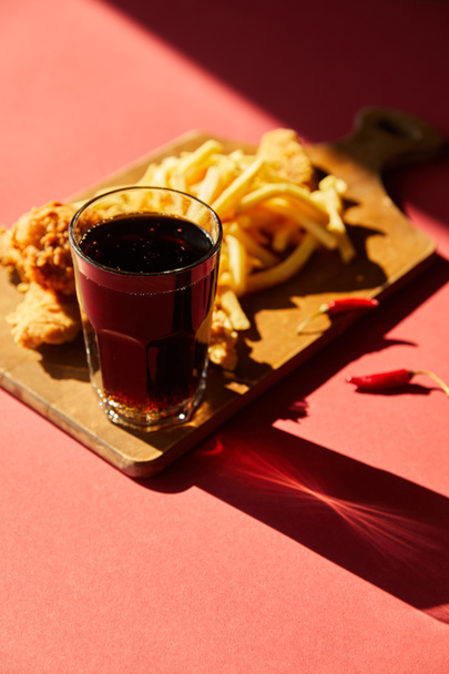 selective focus of spicy deep fried chicken and french fries served on wooden cutting board with soda in sunlight - Photo, Image