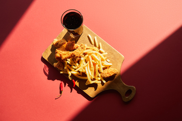 top view of spicy deep fried chicken and french fries served on wooden cutting board with soda in sunlight - 写真・画像