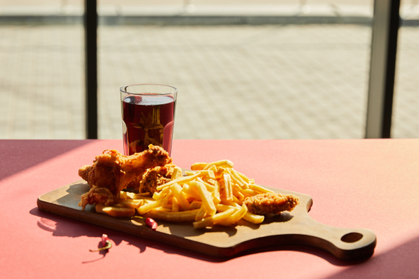 spicy deep fried chicken and french fries served on wooden cutting board with soda in sunlight near window - Foto, Bild