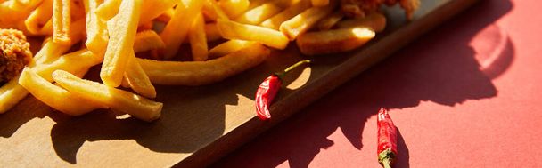 spicy french fries served on wooden cutting board with chili peppers in sunlight, panoramic shot - Фото, зображення