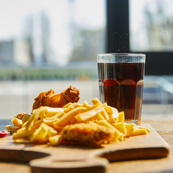 selective focus of deep fried chicken, french fries on board with soda in glass on wooden table in sunlight near window - Photo, Image