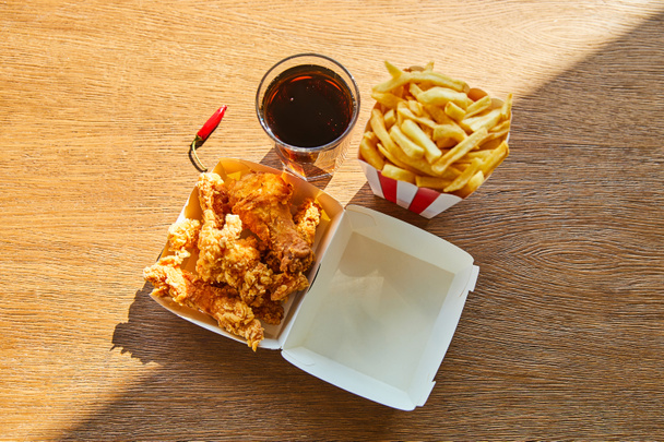 vista superior de pollo frito, papas fritas y refrescos en vidrio sobre mesa de madera a la luz del sol
 - Foto, Imagen