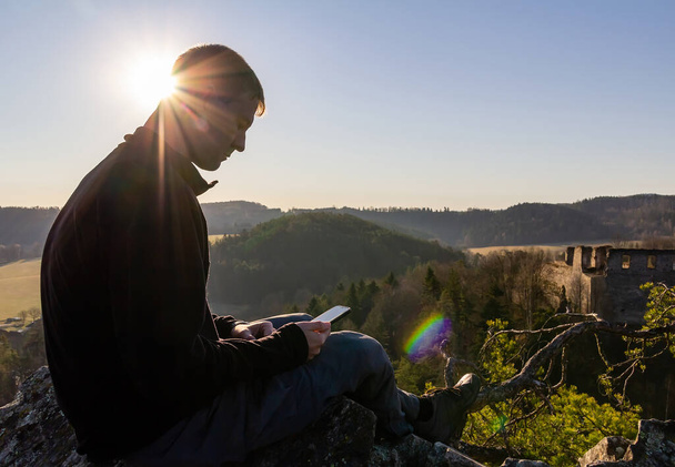 Junger kaukasischer Mann sitzt mit seinem Handy auf einem Felsen mit Ruinen und Sonne. Tschechische Republik - Foto, Bild