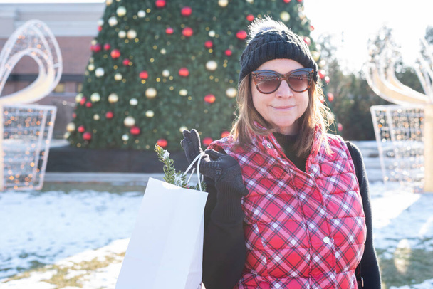 mujer feliz de mediana edad sosteniendo bolsas de compras frente al árbol de Navidad en el centro comercial al aire libre en temporada de fiestas
 - Foto, imagen