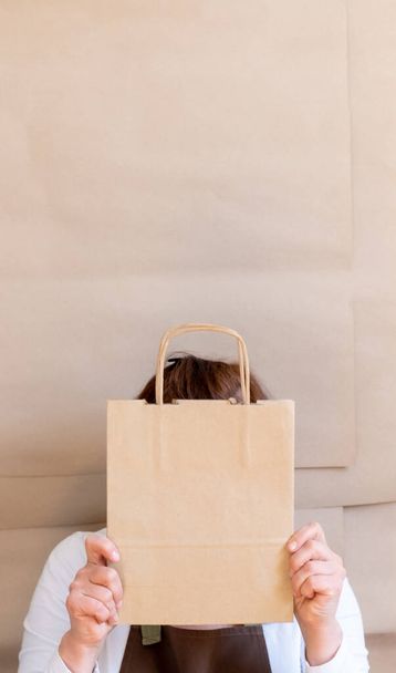 Τα χέρια του Packer κρατάνε μια χάρτινη τσάντα και ένα κουτί. Εργαζόμενος ντυμένος με καφέ ποδιά Συσκευασία Παραγγελία για Πελάτη. - Φωτογραφία, εικόνα