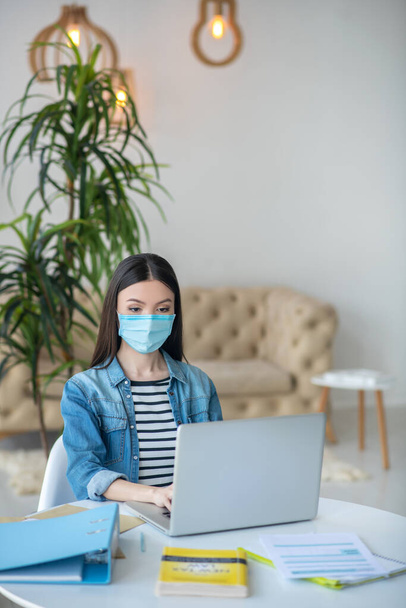 Dark-haired female sitting at her desk, typing on laptop, wearing protective mask - Photo, Image