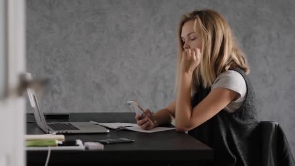 Νεαρή γυναίκα κάθεται σε ένα γραφείο, online μάθηση, φορητό υπολογιστή, και smartphone - Πλάνα, βίντεο