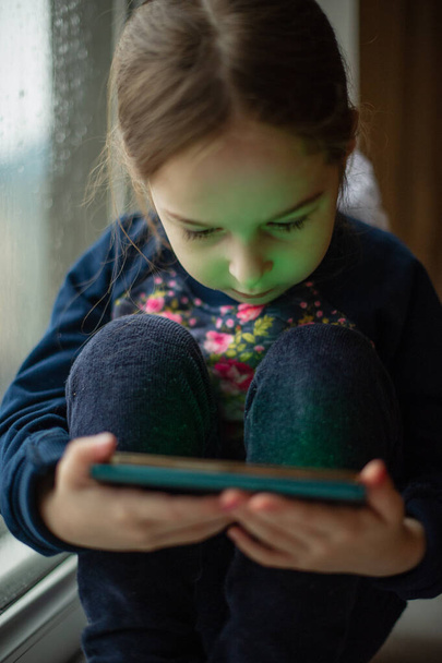 Όμορφο χαριτωμένο Καυκάσιο θηλυκό παιδί κάθεται στο περβάζι του παραθύρου. Ένα κοριτσάκι κάθεται σε ένα περβάζι του παραθύρου και κοιτάζει σε ένα smartphone. Πορτρέτο της σύγχρονης κορίτσι παιδιά κάθεται στο περβάζι με το smartphone - Φωτογραφία, εικόνα
