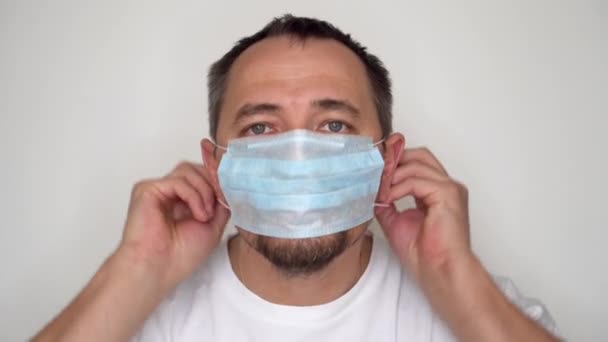A man with a beard is putting on a protective mask. Portrait of an overgrown and unshaven man in a medical mask and the inscription Quarantine on the mask. The isolation and quarantine concept. - Imágenes, Vídeo