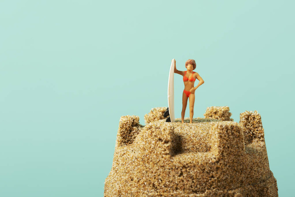 primer plano de una mujer en miniatura en traje de baño, de pie junto a una tabla de surf, en la parte superior de un castillo de arena, sobre un fondo azul con un poco de espacio en blanco a la izquierda
 - Foto, Imagen