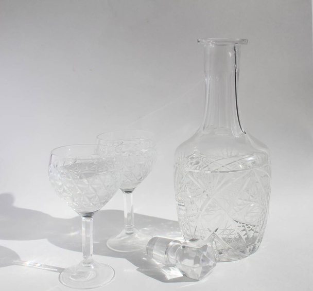 dos vasos de cristal con un patrón de gafas en una pierna delgada con vodka y un decantador con vodka sobre un fondo blanco claro o gris
 - Foto, Imagen