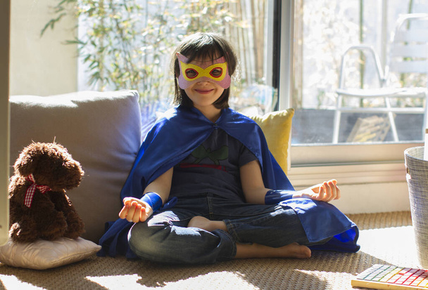 smiling superhero child with mask and cape enjoying happy yoga, fun meditation and mindfulness to relax facing virus lockdown - Fotoğraf, Görsel
