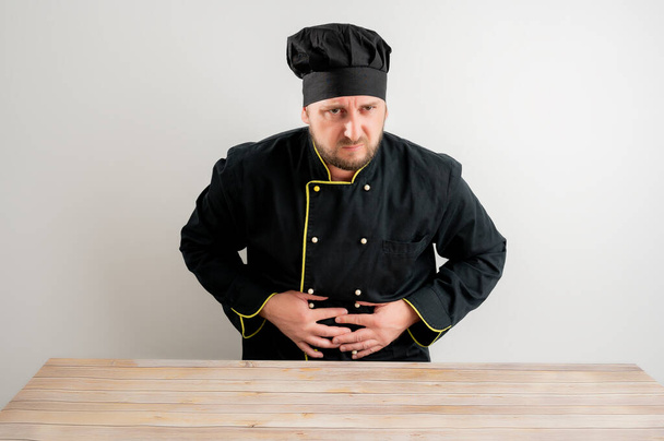 Giovane chef maschio in uniforme nera ha mal di stomaco in posa su uno sfondo bianco isolato
 - Foto, immagini