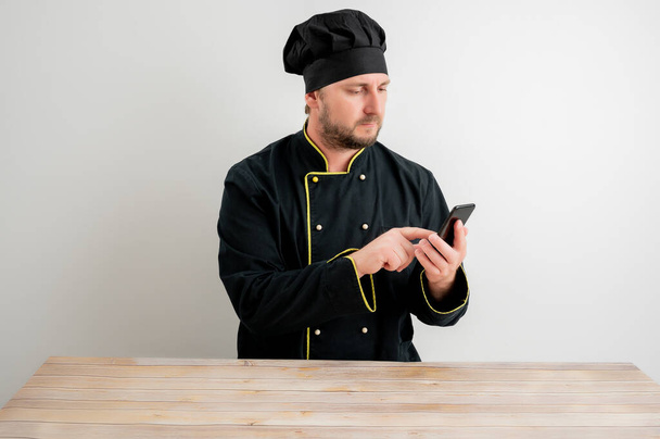 Portrait of young male chef in black uniform with blonde hair, holding smartphone, texting messages posing on a white isolated background - Foto, Imagen