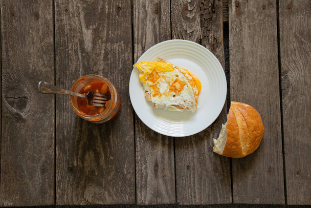ovo frito em uma chapa branca com pão e um jarro de feijão em uma mesa de madeira
 - Foto, Imagem