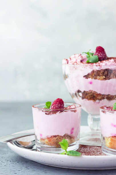 Pink layered dessert trifle, tiramisu or parfait with raspberries and mint decorated with cocoa powder in glasses. No bake summer dessert. Grey concrete background.  - Foto, immagini