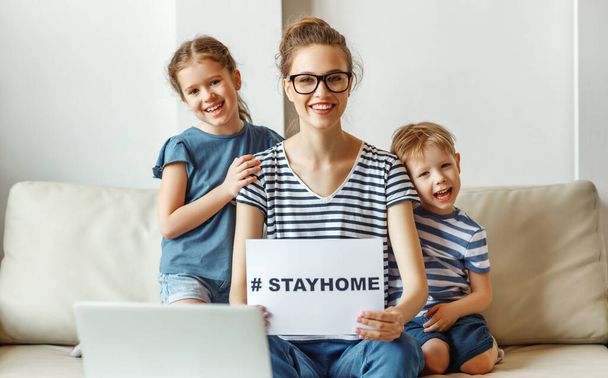Jovencita alegre con niños pequeños sentados frente a la computadora portátil en el sofá y papel de demostración con etiqueta Stay Home para la prevención del coronavirus
 - Foto, imagen