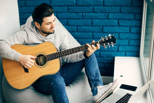 musician playing on fresh new guitar at home with laptop, happy guitarist practicing with bue bricks background - Photo, Image