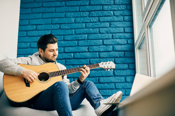 Мужчина мужественности, держащий гитару и сидящий в современной комнате с синей стеной кирпичей и ногой на подоконнике
 - Фото, изображение