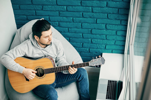 guitarist in casual wear in modern room with blue brick practicing with laptop on the window sill, copy space - Photo, Image