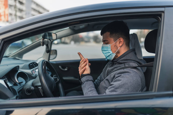 Bir adam arabaya oturur ve ellerini antiseptik jelle yıkar. Arabada sağlık hizmeti kavramı. Maske yüzünde beyazdır. Koronavirüs, hastalık, enfeksiyon, karantina, covid-19 - Fotoğraf, Görsel