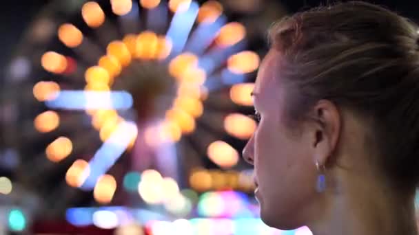 Девушка с длинными светлыми волосами, собранными в хвостик, смотрит на большое колесо обозрения, катается ночью на фоне города. Парк аттракционов, лунный парк. Задний вид 4K видео
. - Кадры, видео