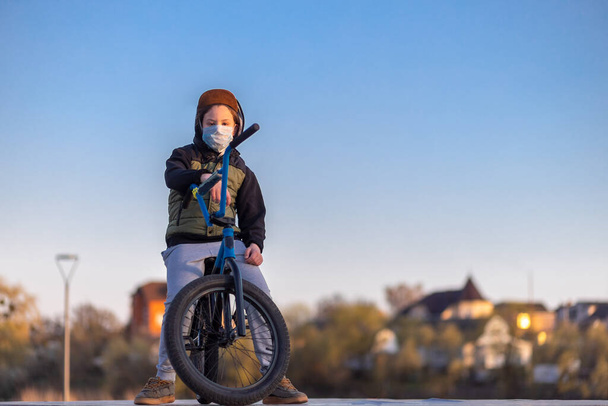 Retrato de un adolescente en la puesta de sol en una máscara en una bicicleta azul. Cuarentena. Está prohibido montar en bicicleta. Covid-19
. - Foto, imagen