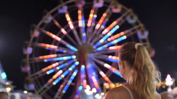 Een meisje met lang blond haar verzameld in een paardenstaart kijkt naar een groot reuzenrad, rijdt tegen de achtergrond van de stad 's nachts. Amusementspark, lunapark. Achteruitkijkspiegel. 4K-video. - Video