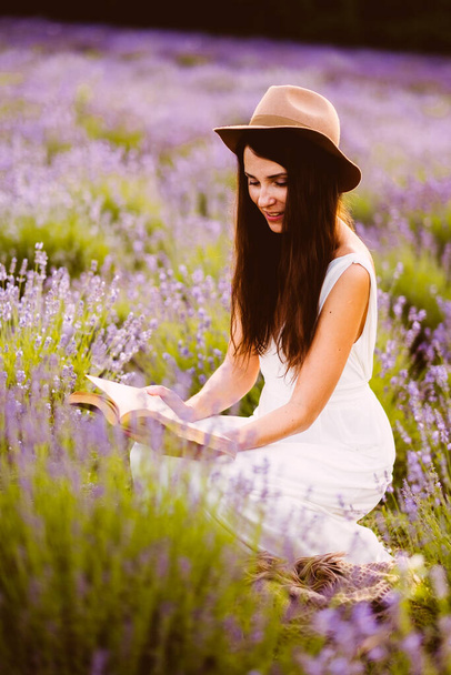 Beyaz elbiseli güzel kadın gün batımında lavanta çiçeklerinin arasında kitap okuyor. - Fotoğraf, Görsel