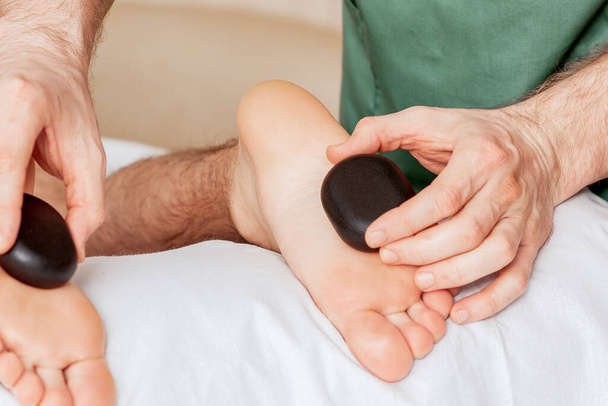 Τα χέρια του θεραπευτή μασάζ δίνοντας μασάζ στα πόδια της γυναίκας από καυτές πέτρες στο σπα. - Φωτογραφία, εικόνα