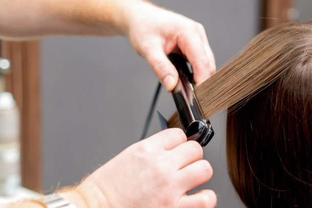 Les mains du coiffeur redressent les cheveux de la femme avec un outil de lissage dans le salon de coiffure, vue arrière
. - Photo, image
