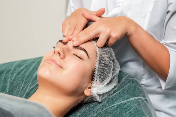 Beautiful woman receiving forehead massage by hands of cosmetologist in beauty salon. - Foto, afbeelding