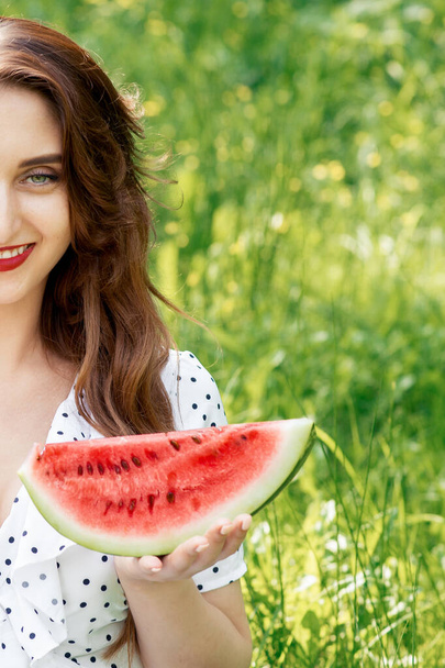 Portrait of half face young woman holding slice of watermelon looking at camera on green grass background. - Fotó, kép