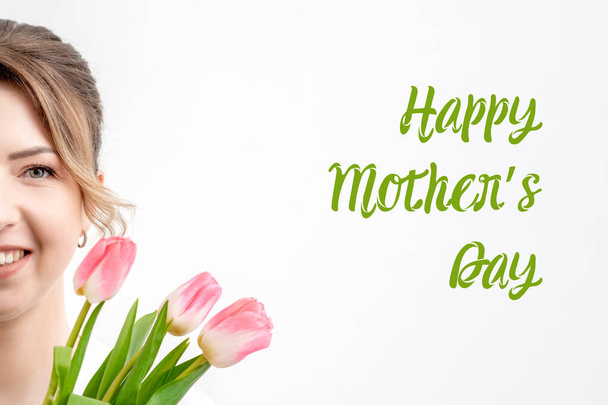 Ευτυχισμένη Ημέρα της Μητέρας πράσινο σημάδι κειμένου και μισό πρόσωπο της νεαρής γυναίκας με μπουκέτο από ροζ τουλίπες άνοιξη σε λευκό φόντο. - Φωτογραφία, εικόνα